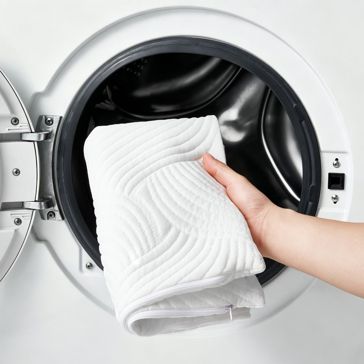 A white quilted fabric pad with a wavy stitched pattern is being held by a hand and lowered into the open drum of a white front-loading washing machine.